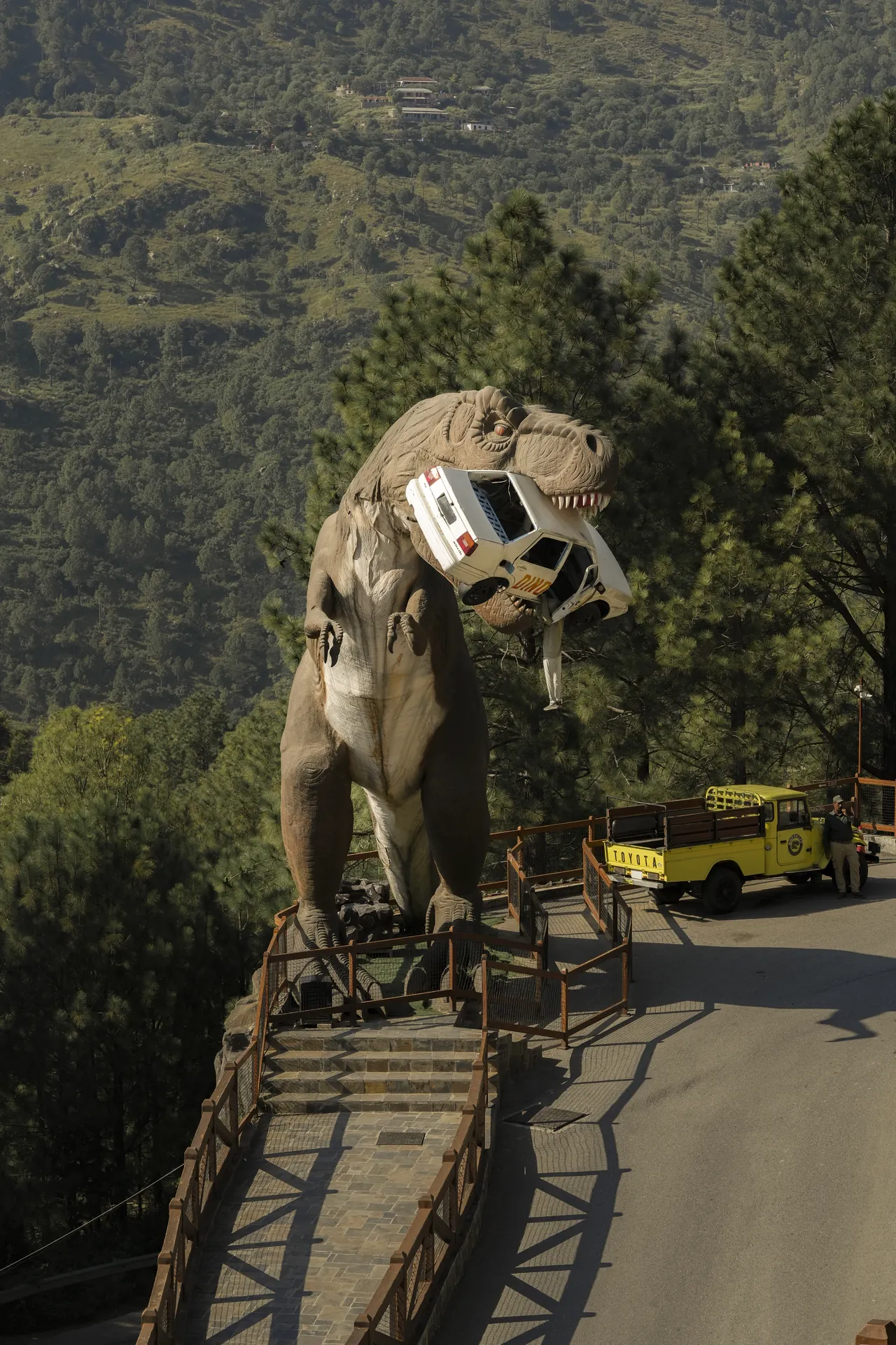 A giant dinosaur standing on a hillside carrying a crumpled jeep in its jaws
